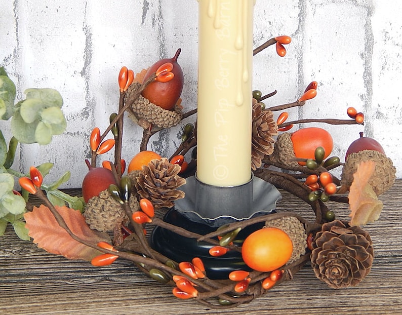 May include: A candle ring with a white candle, brown pine cones, orange and brown acorns, and orange berries. The candle ring is made of brown twigs and is sitting on a wooden surface.