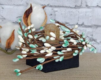 May include: A decorative candle ring featuring brown stems with white and teal-colored berries. The ring sits on a black base, with a small, neutral-colored candle in the center. Cotton bolls are visible in the background.