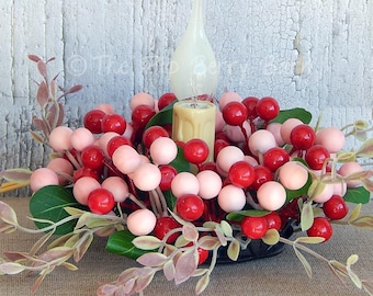 May include: A decorative centerpiece featuring a candle, surrounded by red and pink berries, green leaves, and delicate sprigs. A white glass vase is behind the candle. The arrangement is set in a black bowl.