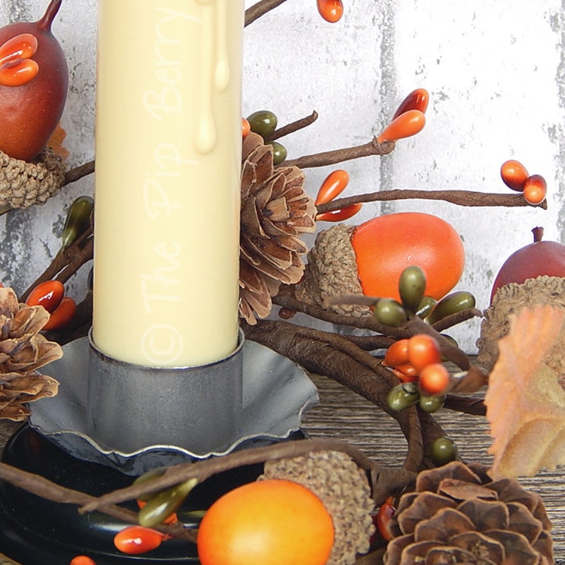 May include: A white pillar candle with a drip of wax sits in a gray metal candle holder. The candle holder is surrounded by a fall-themed wreath with brown pinecones, acorns, and orange berries.