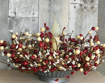 May include: A decorative centerpiece featuring a candle surrounded by a wreath of red, cream, and white berries on brown stems. The berries are arranged in a galvanized metal container, creating a rustic aesthetic.