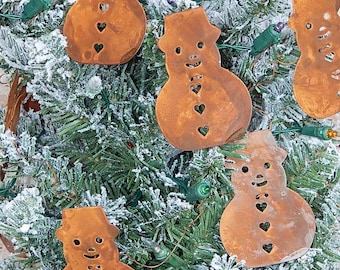 May include: Brown metal snowman ornaments with heart-shaped cutouts are displayed on a snow-covered Christmas tree. The ornaments have a rustic appearance and are illuminated by green Christmas lights.