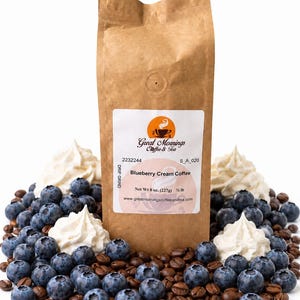 Great Mornings Blueberry Cream Coffee in a kraft paper bag on a white studio background, surrounded by fresh blueberries, dollops of whipped cream, and roasted coffee beans.