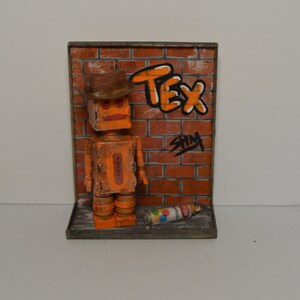 May include: Orange robot wearing a brown cowboy hat standing in front of a brick wall with graffiti that says "TEX" and "SHIM". The robot is holding a spray paint can.