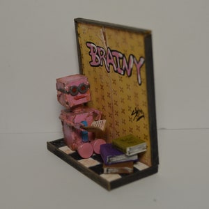 May include: A pink robot with red glasses sits on a checkered floor reading a book. The robot is in front of a wall with the word "BRAINY" written on it in pink spray paint.