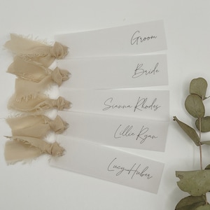 May include: Five white place cards with calligraphy names, including "Groom", "Bride", "Sianna Rhodes", "Lillie Ryan", and "Lucy Huber". The cards are tied with beige frayed ribbon.