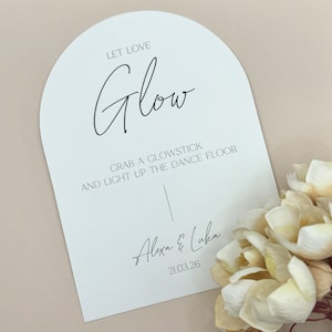 May include: White arch-shaped card with black text that reads "Let Love Glow Grab a Glowstick and Light Up the Dance Floor Alexa & Luka 21.03.26"