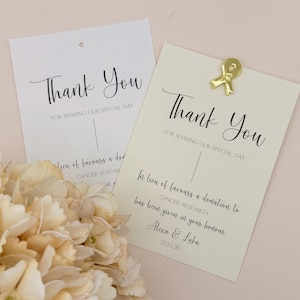 以下が含まれることがあります： 「Thank You」と「For Sharing Our Special Day」という言葉がエレガントな筆記体で書かれた、クリーム色と白の2枚のサンキューカード。クリーム色のカードには金のリボンクリップが付いています。カードには、がん研究への寄付について記載されています。