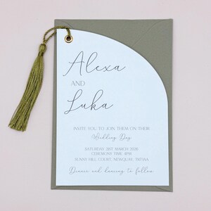 May include: A white wedding invitation with a green tassel attached. The invitation is for Alexa and Luka's wedding on Saturday, March 21st, 2026 at 4pm at Sunny Hill Court, Newquay, TR7 1AA. The invitation reads "Invite you to join them on their Wedding Day".