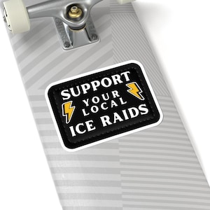 Puede incluir: Una pegatina negra con texto blanco que dice "SUPPORT YOUR LOCAL ICE RAIDS" y gráficos de rayos amarillos. La pegatina está en una tabla de skate con un camión plateado.