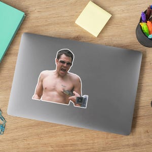 May include: A sticker featuring a shirtless man with a surprised expression, wearing sunglasses and holding a small alligator in his hand. The man is looking at the camera.