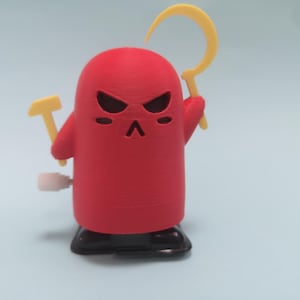 Op de afbeelding: Een rode opwindbare speelgoedfiguur met een strenge uitdrukking, die een gele hamer en sikkel vasthoudt. Het speelgoed heeft zwarte ogen en een zwarte basis. De opwindsleutel is zichtbaar.