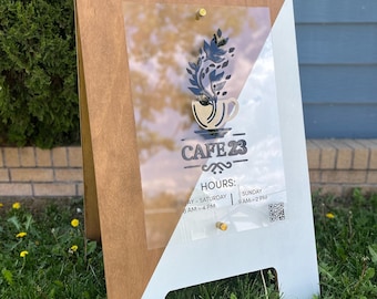 May include: A-frame sign for Cafe 23, featuring a clear acrylic panel with a coffee cup and leaf design. The sign has a wooden frame and white base, with hours of operation and a QR code. It is displayed on a grassy lawn.