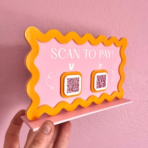 May include: A pink and orange sign with the text "SCAN TO PAY!" and two QR codes. The sign is on a stand and is being held by a hand.