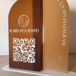 May include: A brown and white sign with the text "Board Housewives" and a QR code. The sign also has the text "Follow us on Instagram" in white on a brown background.