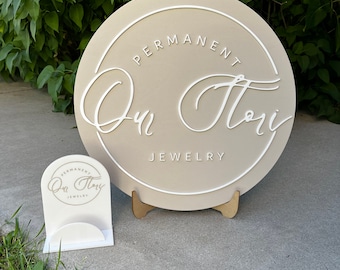 Könnte beinhalten: Ein rundes, beiges Schild mit weißer Aufschrift "PERMANENT Our Story JEWELRY". Das Schild steht auf einem Holzständer. Eine kleinere Version des Schildes ist ebenfalls zu sehen.