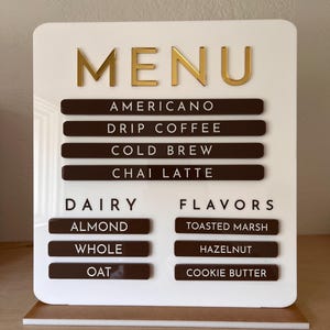 May include: White menu board with gold lettering that reads "MENU". The board lists coffee options: Americano, Drip Coffee, Cold Brew, Chai Latte. It also lists dairy options: Almond, Whole, Oat. Flavors include: Toasted Marsh, Hazelnut, Cookie Butter.