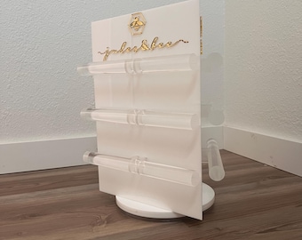 May include: A white, rotating jewelry display stand with clear acrylic rods for hanging items. The stand features gold lettering and a bee logo. The display is on a white base and is set against a neutral wall and wood-look flooring.