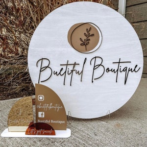 May include: A white wooden sign with the words "Buetiful Boutique" in black script. The sign has a brown circle with a leaf design. A smaller sign displays social media handles and the phrase "Let's get social!"