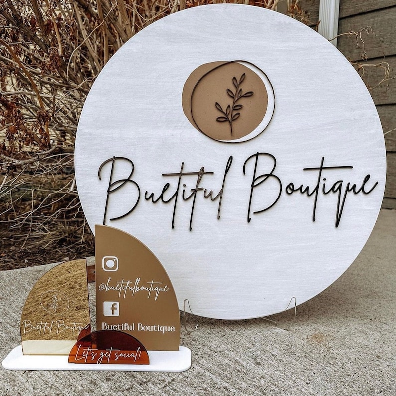 May include: A white wooden sign with the words "Buetiful Boutique" in black script. The sign has a brown circle with a leaf design. A smaller sign displays social media handles and the phrase "Let's get social!"