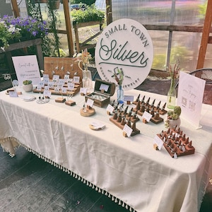 May include: A display of handcrafted jewelry at an outdoor market. The table is covered with a cream-colored tablecloth and features earrings, rings, and other silver jewelry. A sign reads "Small Town Silvers, Handcrafted Jewelry."