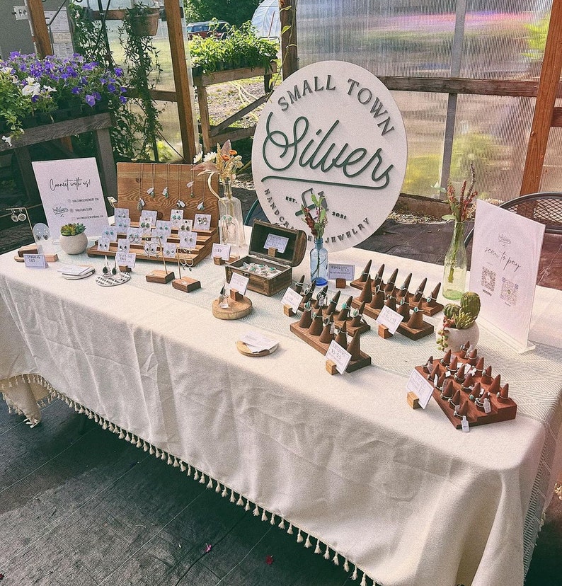 May include: A display of handcrafted jewelry at an outdoor market. The table is covered with a cream-colored tablecloth and features earrings, rings, and other silver jewelry. A sign reads "Small Town Silvers, Handcrafted Jewelry."