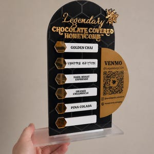 May include: A black and gold sign with the text "Legendary Chocolate Covered Honeycomb." The sign lists flavors: Golden Chai, Vampire Edition, Dark Roast Espresso, Orange Creamsicle, and Pina Colada. A QR code and social media handles are also displayed.