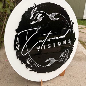 May include: A round, white sign with a black center featuring the text "Entwined Visions" and two koi fish illustrations. The sign also includes "EST. 2024" in white lettering. The sign is displayed on a stand.