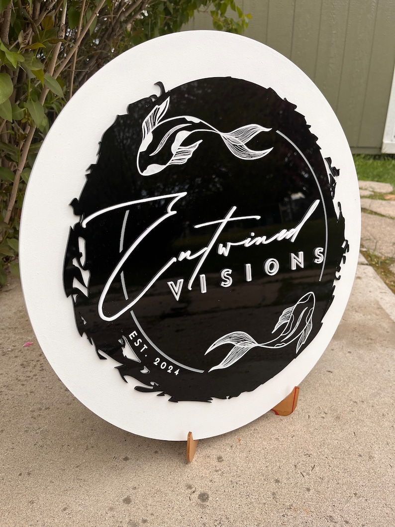 May include: A round, white sign with a black center featuring the text "Entwined Visions" and two koi fish illustrations. The sign also includes "EST. 2024" in white lettering. The sign is displayed on a stand.