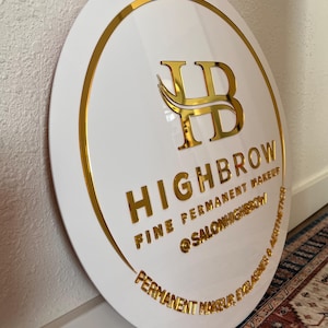 May include: A white, round sign with gold lettering and trim. The sign reads "HIGHBROW FINE PERMANENT MAKEUP @SALONHIGHBROW PERMANENT MAKEUP, EYELASHES & AESTHETICS". The logo features gold intertwined letters "HB".