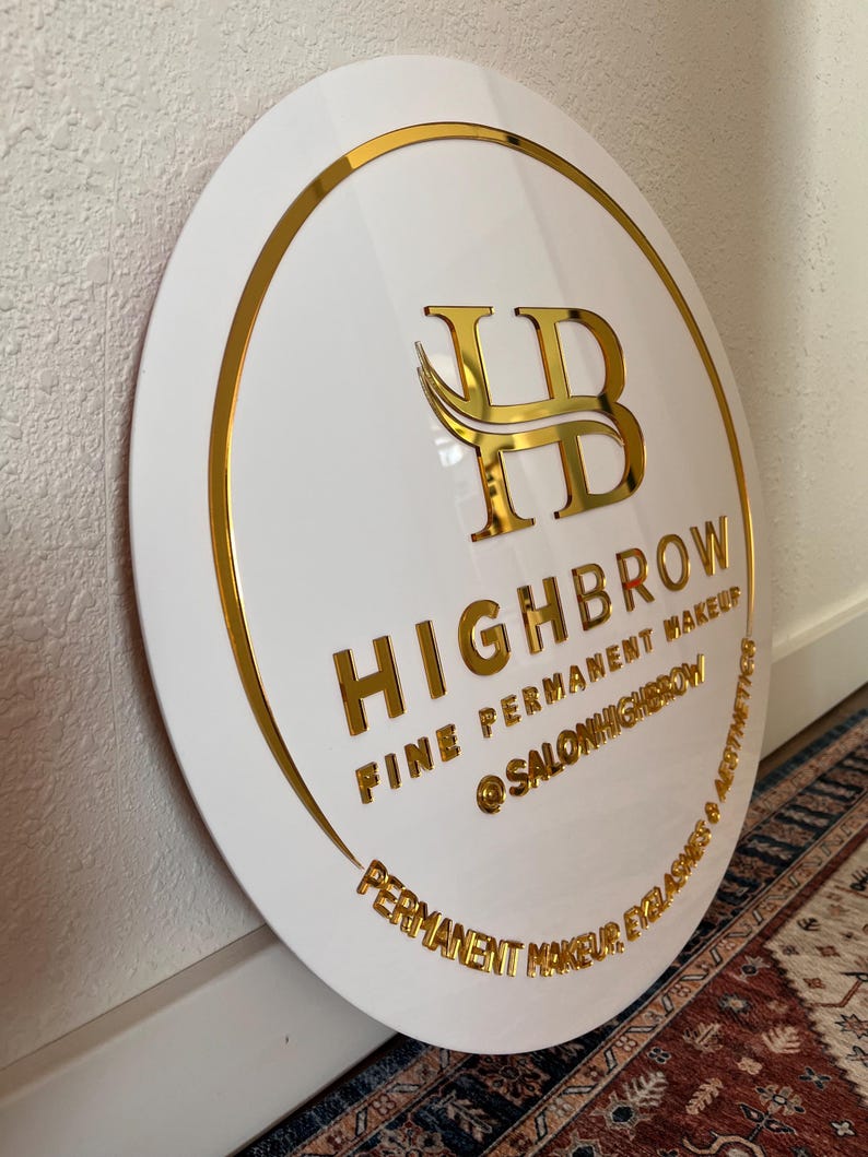 May include: A white, round sign with gold lettering and trim. The sign reads "HIGHBROW FINE PERMANENT MAKEUP @SALONHIGHBROW PERMANENT MAKEUP, EYELASHES & AESTHETICS". The logo features gold intertwined letters "HB".