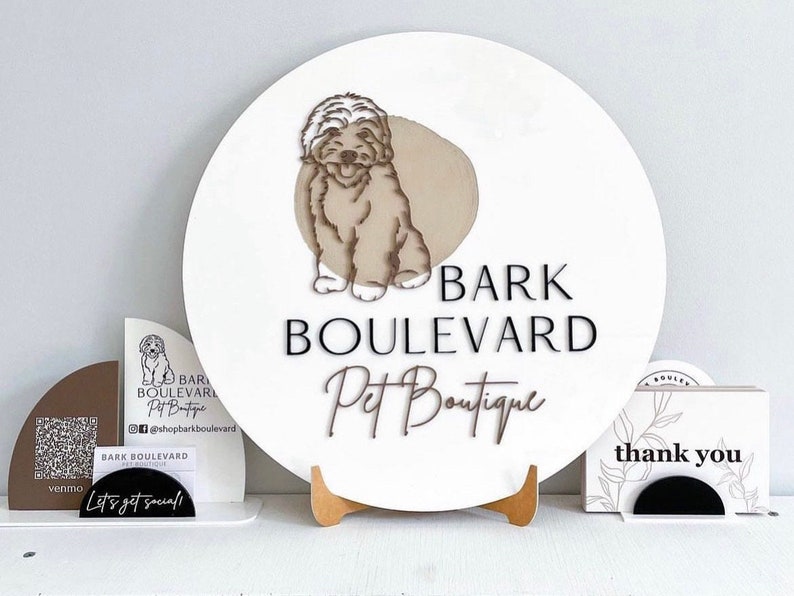May include: A white, round sign with the text "BARK BOULEVARD Pet Boutique" and a dog illustration. Additional signage includes a QR code, social media handles, and "thank you" cards. The sign is displayed on a wooden stand.