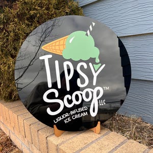 May include: A round black sign with the text "Tipsy Scoop" and an ice cream cone graphic. The ice cream is green, and the cone is tan. The sign is propped up on a small wooden stand.