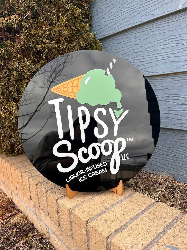 May include: A round black sign with the text "Tipsy Scoop" and an ice cream cone graphic. The ice cream is green, and the cone is tan. The sign is propped up on a small wooden stand.