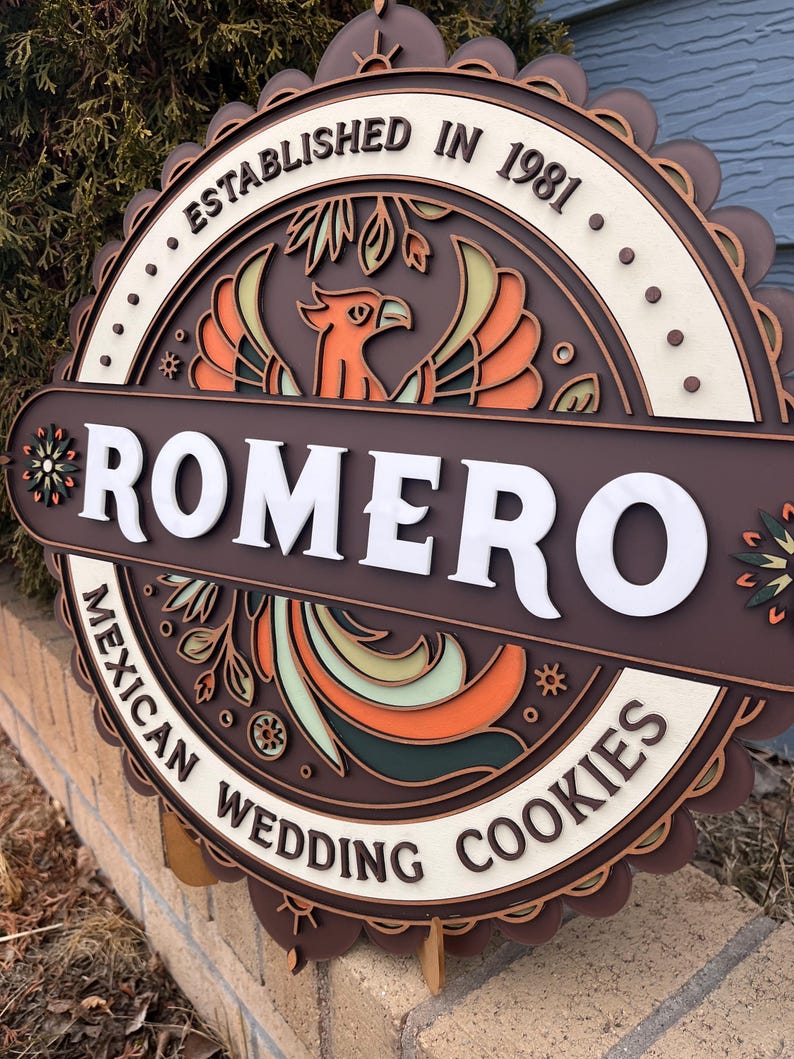 May include: A layered wooden sign with the name "ROMERO" in white block letters. The sign features a phoenix design in orange, green, and brown, with the text "ESTABLISHED IN 1981" and "MEXICAN WEDDING COOKIES".