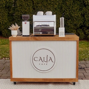 May include: A white coffee cart with a wooden top and bottom. The cart has a white sign with the words "Calia Cafe" in a circle. There is a coffee machine, grinder, and a glass jar on the counter.