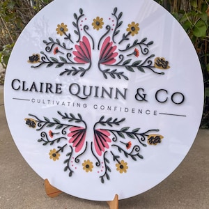 May include: A round white sign with the text "CLAIRE QUINN & CO" and "CULTIVATING CONFIDENCE". The sign features a floral design with pink, yellow, and green accents. The sign is displayed on a wooden stand.