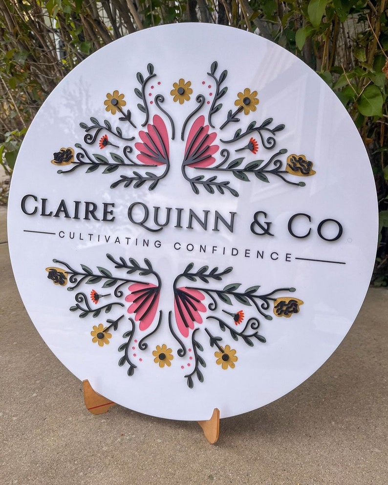 May include: A round white sign with the text "CLAIRE QUINN & CO" and "CULTIVATING CONFIDENCE". The sign features a floral design with pink, yellow, and green accents. The sign is displayed on a wooden stand.