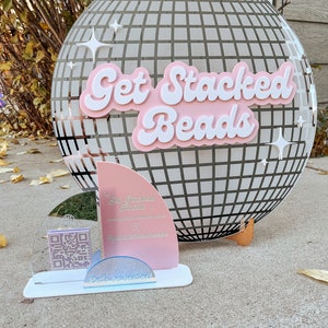 May include: A pink and white acrylic sign with the text "Get Stacked Beads" in a retro font. The sign is in front of a silver disco ball with a grid pattern. The sign also includes a QR code and the text "Get Stacked Beads" and "@getstackedbeads".