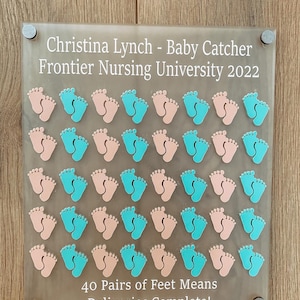 May include: A clear acrylic plaque with the text "Christina Lynch - Baby Catcher Frontier Nursing University 2022" and "40 Pairs of Feet Means Deliveries Complete!"  The plaque has 40 pairs of baby footprints, alternating between pink and turquoise.