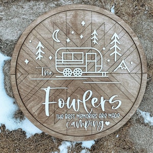 May include: A round wooden sign with a white camper, trees, stars, and a crescent moon. The text "The Fowlers The Best Memories Are Made Camping" is written in white on the sign.