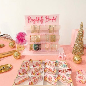 May include: A pink display stand with the text "Brightfully Beaded" in pink lettering. The stand is filled with colorful beaded bracelets. The bracelets are displayed on a white background. The stand is on a pink table with a gold tray of bracelets in front of it.