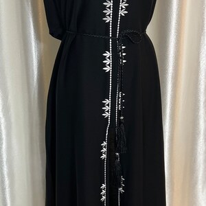 Op de afbeelding: Zwarte maxi-jurk met witte bloemenborduursels langs de voorkant en de halslijn. De jurk heeft een V-hals, lange mouwen en een bijpassende zwarte riem. De jurk wordt op een mannequin getoond.