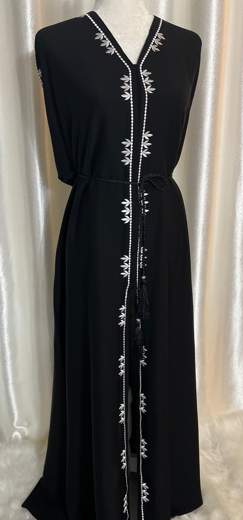 Op de afbeelding: Zwarte maxi-jurk met witte bloemenborduursels langs de voorkant en de halslijn. De jurk heeft een V-hals, lange mouwen en een bijpassende zwarte riem. De jurk wordt op een mannequin getoond.