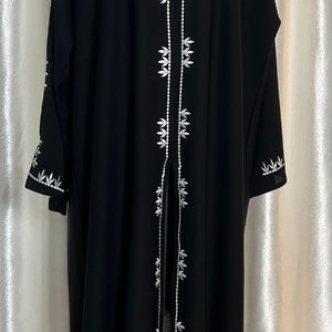 Op de afbeelding: Zwarte abaya met witte bloemenborduurwerk aan de voorkant en mouwen. Het kledingstuk met lange mouwen heeft een V-hals en wordt op een mannequin gepresenteerd. Het borduurwerk is gedetailleerd en symmetrisch.