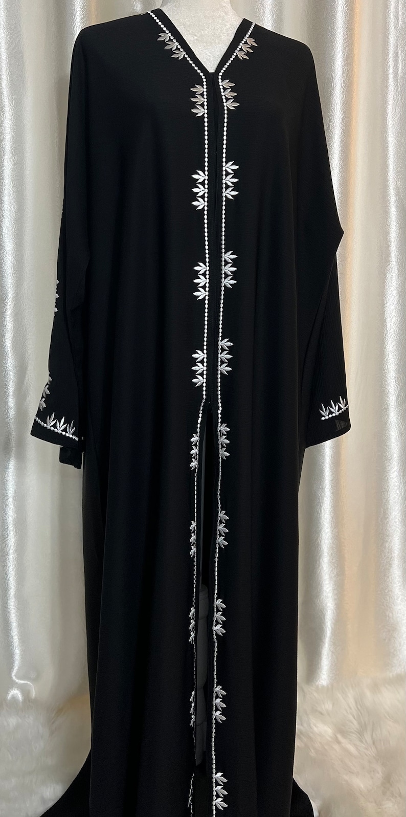 Op de afbeelding: Zwarte abaya met witte bloemenborduurwerk aan de voorkant en mouwen. Het kledingstuk met lange mouwen heeft een V-hals en wordt op een mannequin gepresenteerd. Het borduurwerk is gedetailleerd en symmetrisch.
