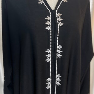 Op de afbeelding: Zwarte abaya met witte bladborduursels langs de halslijn en de voorkant. Het kledingstuk met lange mouwen heeft gedetailleerde witte borduursels op de manchetten. De abaya wordt op een mannequin gepresenteerd en toont het vloeiende ontwerp en de elegante details.