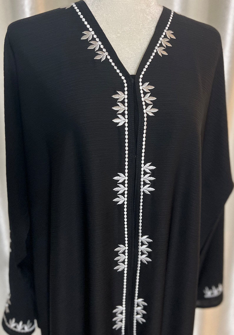 Op de afbeelding: Zwarte abaya met witte bladborduursels langs de halslijn en de voorkant. Het kledingstuk met lange mouwen heeft gedetailleerde witte borduursels op de manchetten. De abaya wordt op een mannequin gepresenteerd en toont het vloeiende ontwerp en de elegante details.
