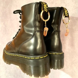 Puede incluir: Botas de plataforma de cuero negro con costuras amarillas y un amuleto dorado sujeto a cada bota. Los amuletos tienen forma de anillo con un cristal rosa colgando. Las botas tienen una correa negra con el texto "Dr. Martens" escrito en blanco.