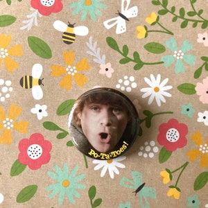 May include: A round button with a picture of a man with a surprised expression. The button has the text "Po-Ta-Toes!" in yellow letters on a black background.